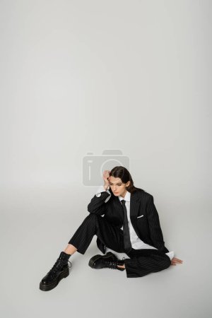pretty woman in laced-up boots and oversize formal wear sitting with hand near head on grey background