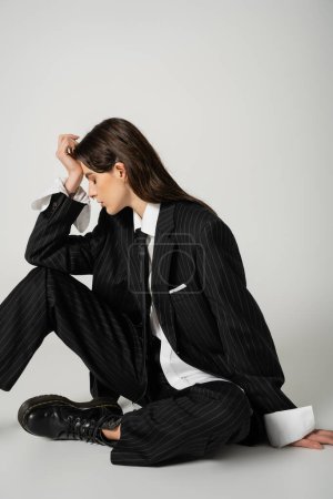 brunette model in black elegant suit and rough footwear sitting with hand near face and closed eyes on grey background