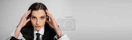 Foto de Portrait of brunette woman in stylish attire touching head while looking at camera isolated on grey, banner - Imagen libre de derechos