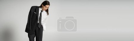 stylish brunette woman in oversize formal wear standing and looking down on grey background, banner