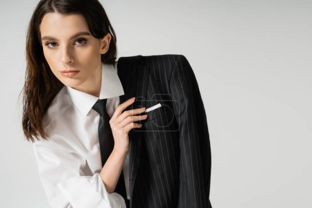 Foto de Stylish woman with makeup and piercing wearing white shirt and holding black blazer isolated on grey - Imagen libre de derechos
