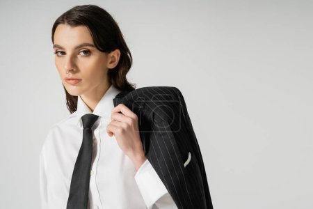 trendy woman in elegant formal wear looking at camera while standing with black blazer isolated on grey