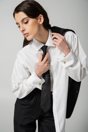 trendy woman in oversize shirt touching black tie while posing isolated on grey
