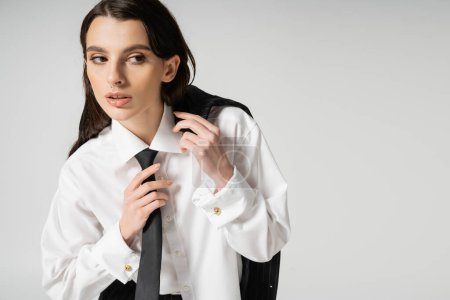 young woman in white shirt touching black tie while holding blazer and looking away isolated on grey