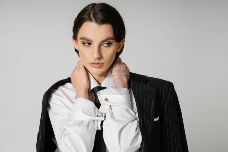 brunette woman in white shirt and black tie posing with blazer and hands on neck isolated on grey