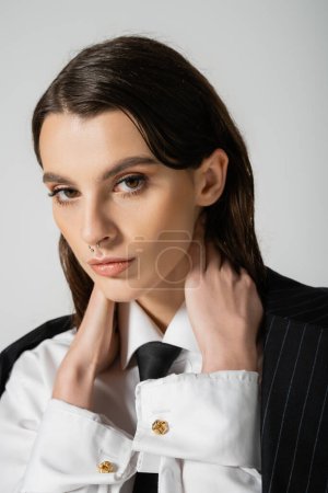 portrait of stylish woman in white shirt and black tie touching neck and looking at camera isolated on grey