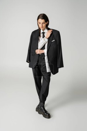full length of young woman in black elegant suit and rough leather boots looking down on grey background