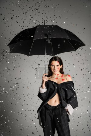 Photo for Cheerful woman with red lip prints on body wearing black clothes and standing with umbrella under falling confetti on grey background - Royalty Free Image