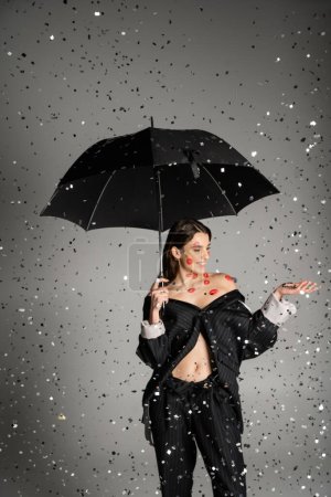 Photo for Happy woman with red kiss prints on body wearing trendy clothes and standing under black umbrella and confetti on grey - Royalty Free Image