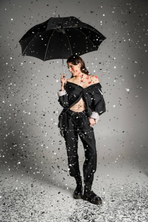 happy woman in black stylish clothes standing with umbrella under sparkling confetti on grey 