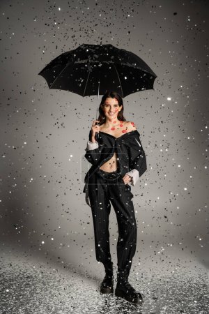 Photo for Happy young woman in stylish clothes standing with black umbrella under shiny confetti on grey background - Royalty Free Image