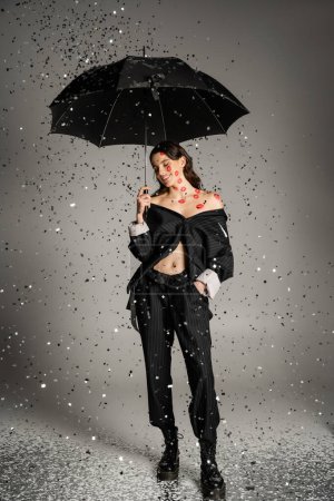 happy and stylish woman with red lip prints on body standing with black umbrella under shiny confetti on grey background