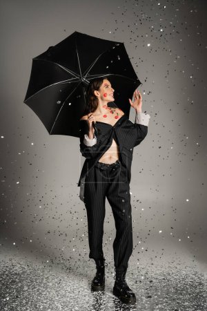 cheerful and trendy woman looking at falling confetti while standing under black umbrella on grey background