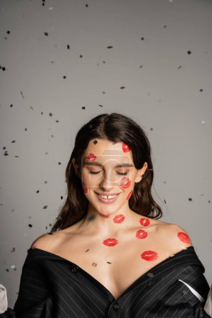 Photo for Brunette woman with red kiss prints on face and bare shoulders smiling with closed eyes on grey background - Royalty Free Image