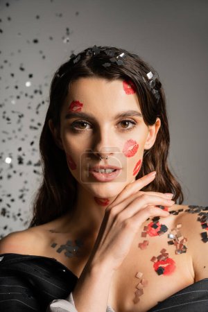 charming woman with red kiss prints on face looking at camera near sparkling confetti on grey 