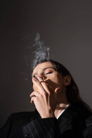 young woman in black and striped blazer smoking with closed eyes isolated on grey with copy space