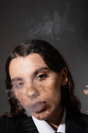 Téléchargez les photos : Portrait of stylish brunette woman smoking and looking at camera isolated on dark grey - en image libre de droit