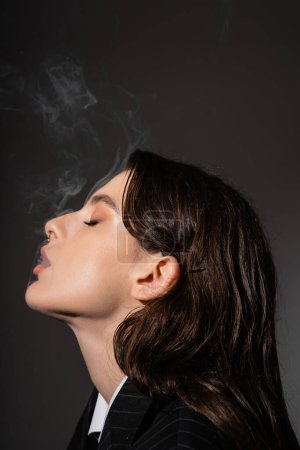 Photo for Profile of stylish brunette woman with makeup and piercing smoking isolated on dark grey - Royalty Free Image