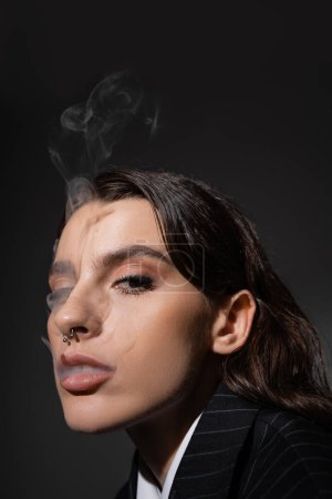 portrait of pretty brunette woman with piercing and makeup smoking and looking at camera isolated on black
