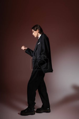 Photo for Side view of young model in black formal wear and rough leather boots posing on brown - Royalty Free Image