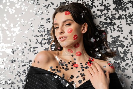 Photo for Top view of stylish woman with red lip prints lying near festive confetti and looking at camera on grey background - Royalty Free Image