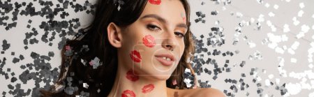 top view of pretty brunette woman with red kiss prints looking at camera near shiny silver confetti on grey background, banner