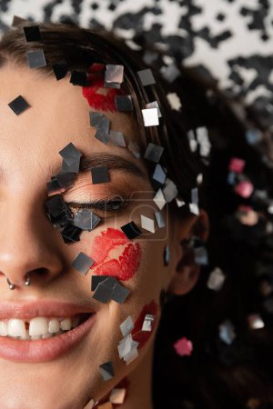 close up portrait of cropped woman with red lipstick marks smiling with closed eyes near sparkling confetti on grey background