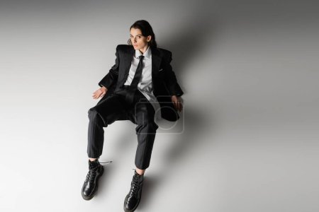 high angle view of brunette woman in black elegant suit and rough leather boots sitting and looking at camera on grey background