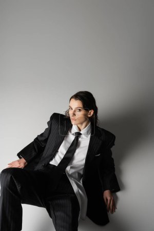 Photo for High angle view of brunette woman in black oversize suit and white shirt sitting and looking at camera on grey background - Royalty Free Image