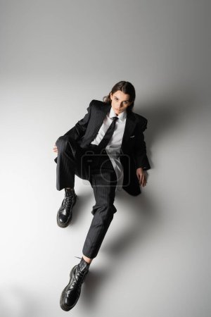 Photo for High angle view of young woman in elegant formal wear and rough boots looking at camera while sitting on grey - Royalty Free Image
