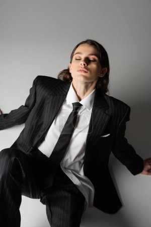 high angle view of sensual woman in white shirt and black striped suit sitting with closed eyes on grey background