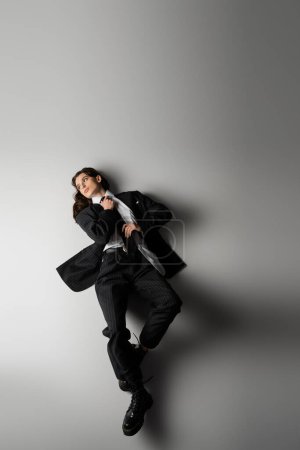 high angle view of model in elegant formal wear and rough boots touching black tie while lying on grey 