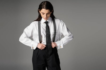 brunette woman in white shirt and black tie adjusting oversize trousers with suspenders isolated on grey