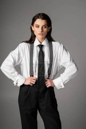 Téléchargez les photos : Brunette woman in white oversize shirt and black tie holding black trousers with suspenders isolated on grey - en image libre de droit