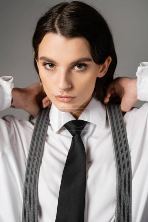 portrait of brunette woman in white shirt and black tie posing with hands near neck isolated on grey