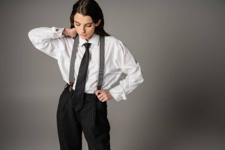 young and stylish woman in oversize shirt and black striped pants touching neck on grey background