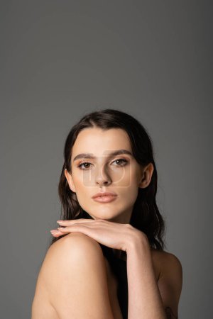 portrait of sexy brunette woman with makeup and piercing posing with hand on bare shoulder isolated on grey