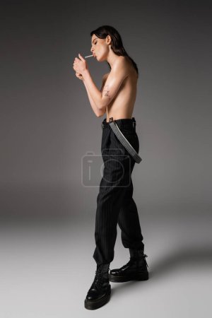 Téléchargez les photos : Full length of shirtless woman in black trousers and rough boots lighting cigarette on grey background - en image libre de droit