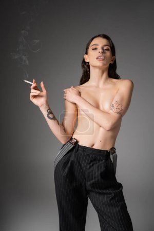 Téléchargez les photos : Young shirtless woman in black striped pants covering bust and looking at camera while smoking on grey background - en image libre de droit
