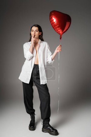 full length of woman in unbuttoned shirt and pants with rough leather boots holding red balloon and smoking on grey 