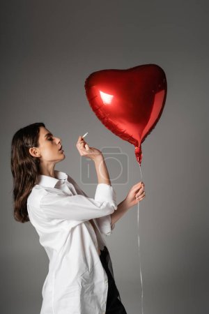 Téléchargez les photos : Side view of brunette woman in white shirt holding cigarette near red heart-shaped balloon on grey - en image libre de droit
