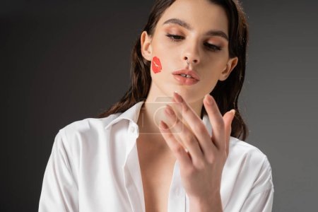 young brunette woman with red kiss print on face looking at hand on grey background