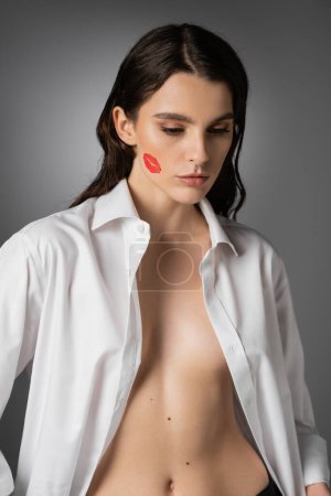 sexy woman with red lip print on face posing in white unbuttoned shirt on grey 