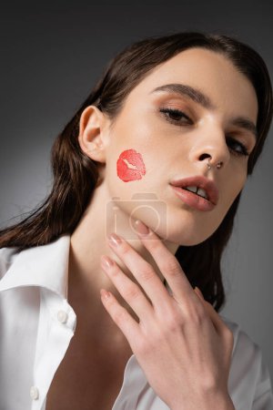 portrait of brunette woman with red kiss print on face touching neck and looking at camera on grey background