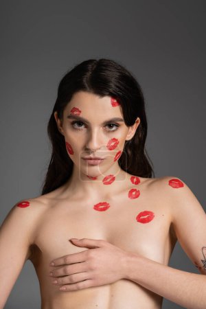 sexy shirtless woman with red kiss prints on body and face covering breast with hand and looking at camera isolated on grey
