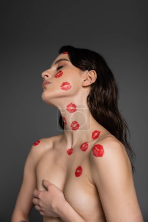 sexy brunette woman with red kiss prints on shirtless body and face covering breast with hand isolated on grey