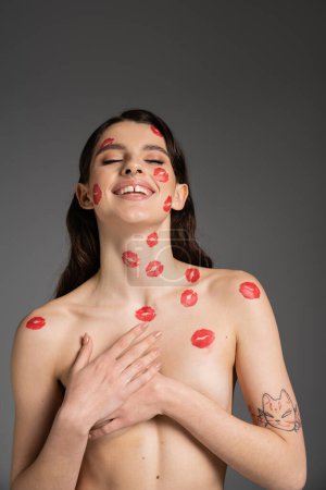 Foto de Joyful shirtless woman with red kisses on face and body covering breast with hands isolated on grey - Imagen libre de derechos