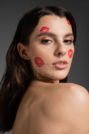 portrait of brunette woman with bare shoulder and red kiss prints looking at camera isolated on grey