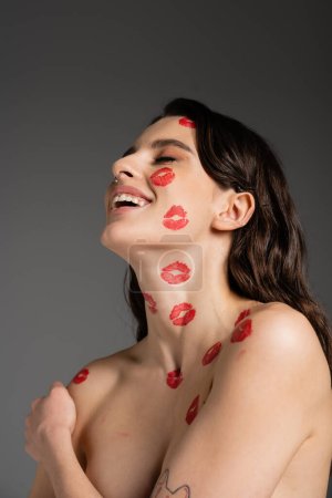 young brunette woman with red kiss prints on face and shirtless body laughing with closed eyes isolated on grey