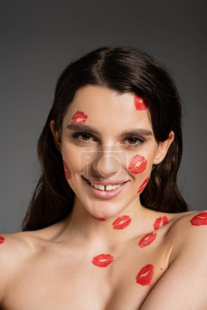 Foto de Young and happy woman with red kiss prints on face and bare shoulders looking at camera isolated on grey - Imagen libre de derechos